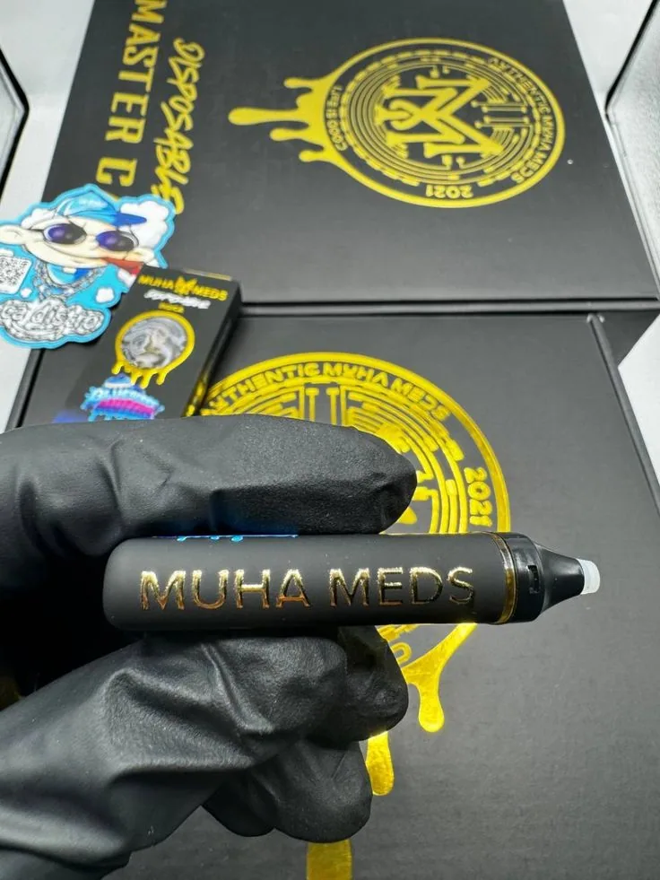 A gloved hand holding a Muha Meds disposable with the small indicator lights visible along the body, held against gold-sealed packaging.