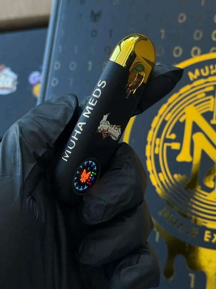 A gloved hand holding a Muha Meds disposable, its circular indicator display lit up in colour against dark branded packaging.